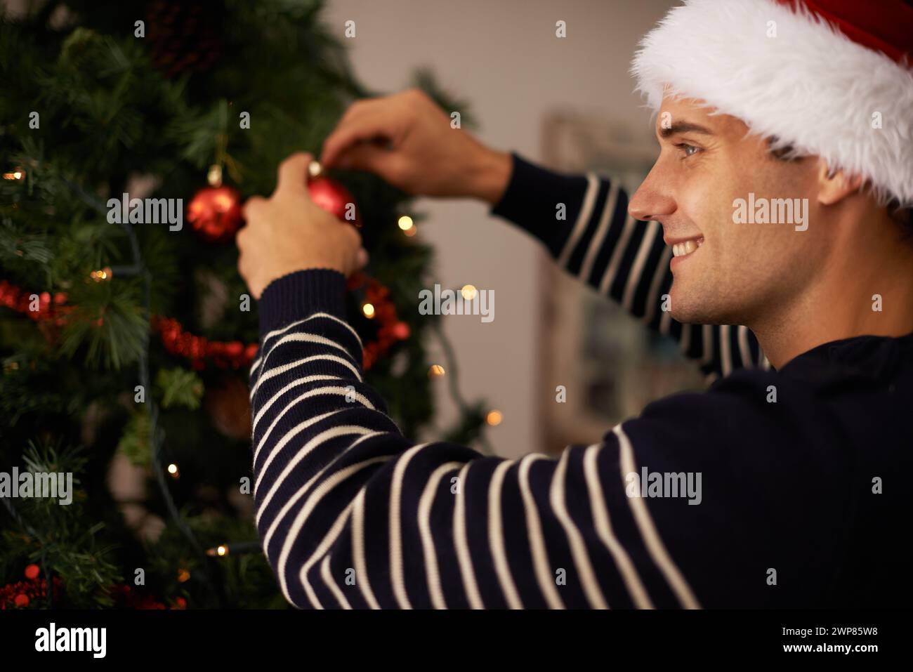 Salon, maison et homme avec le sourire pour les décorations sur arbre pour la fête de Noël ou la célébration. Appartement, bonheur et personne masculine avec joie dans Banque D'Images