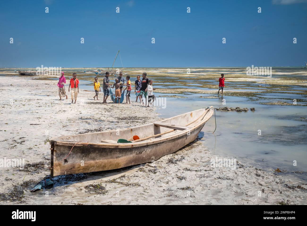 Un canoë en bois à côté des enfants pêchant en utilisant une vieille bâche dans des bassins de marée à marée basse sur la plage de Jambiani, Zanzibar, Tanzanie Banque D'Images