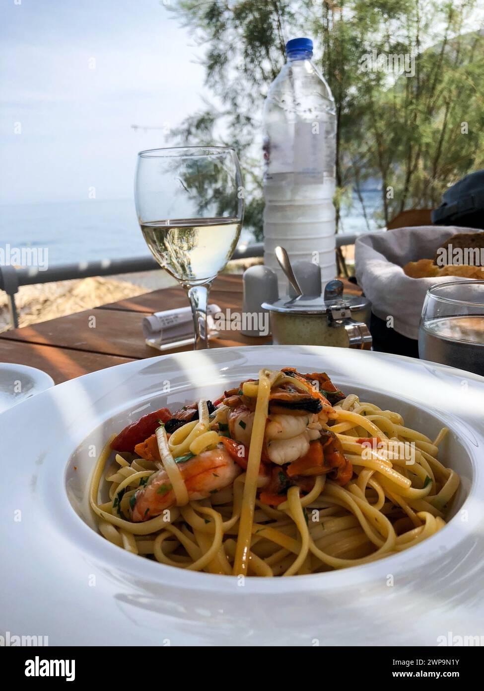 Un délicieux repas composé de pâtes de fruits de mer et de vin blanc servi en plein air, avec un magnifique décor de bord de mer créant une ambiance culinaire parfaite. Banque D'Images