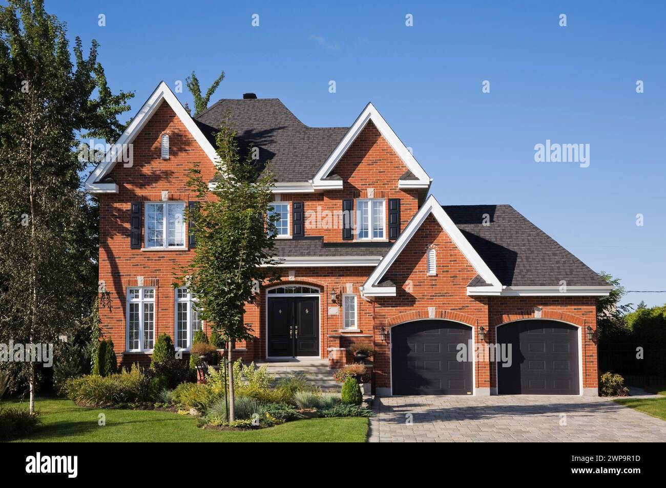 Maison haut de gamme de deux étages en briques rouges et noires avec garage pour deux voitures, allée en pierre pavée et cour avant paysagée en été. Banque D'Images