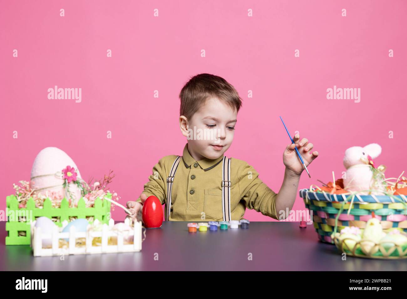 Charmant jeune avec un sourire fabriquant des œufs et des décorations pour les festivités de Pâques, en utilisant des fournitures d'art et de la peinture à l'aquarelle. Petit garçon heureux se sentant excité par l'activité de peinture. Banque D'Images