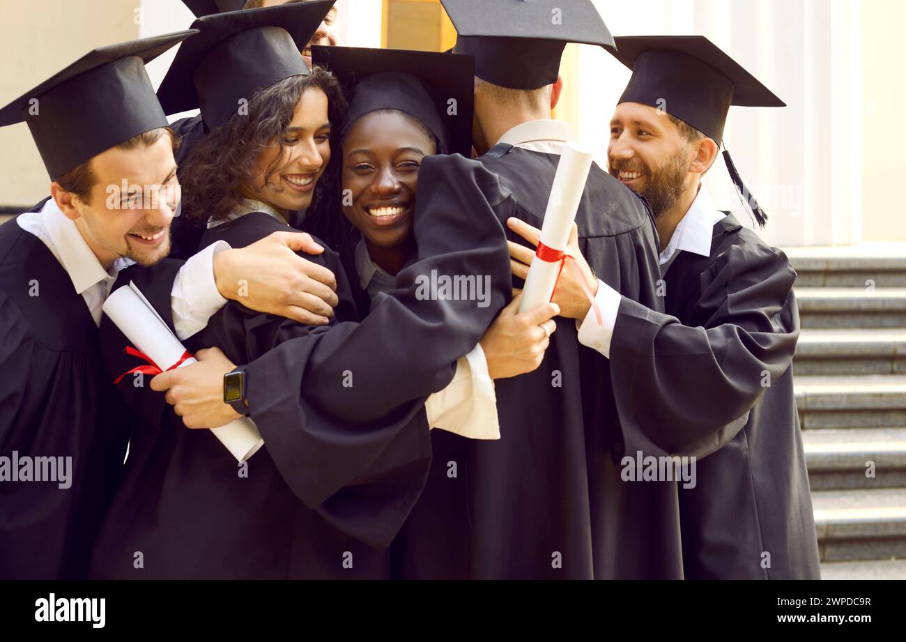 Groupe d'étudiants heureux et divers dans des casquettes et des robes de soirée s'embrassent les uns les autres le jour de la remise des diplômes Banque D'Images