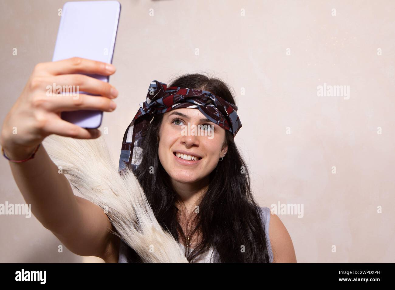 Une jeune femme animée ornée d'un foulard élégant sourit alors qu'elle capture un selfie à l'aide de son smartphone, dégageant un sentiment de joie et de modernité Banque D'Images