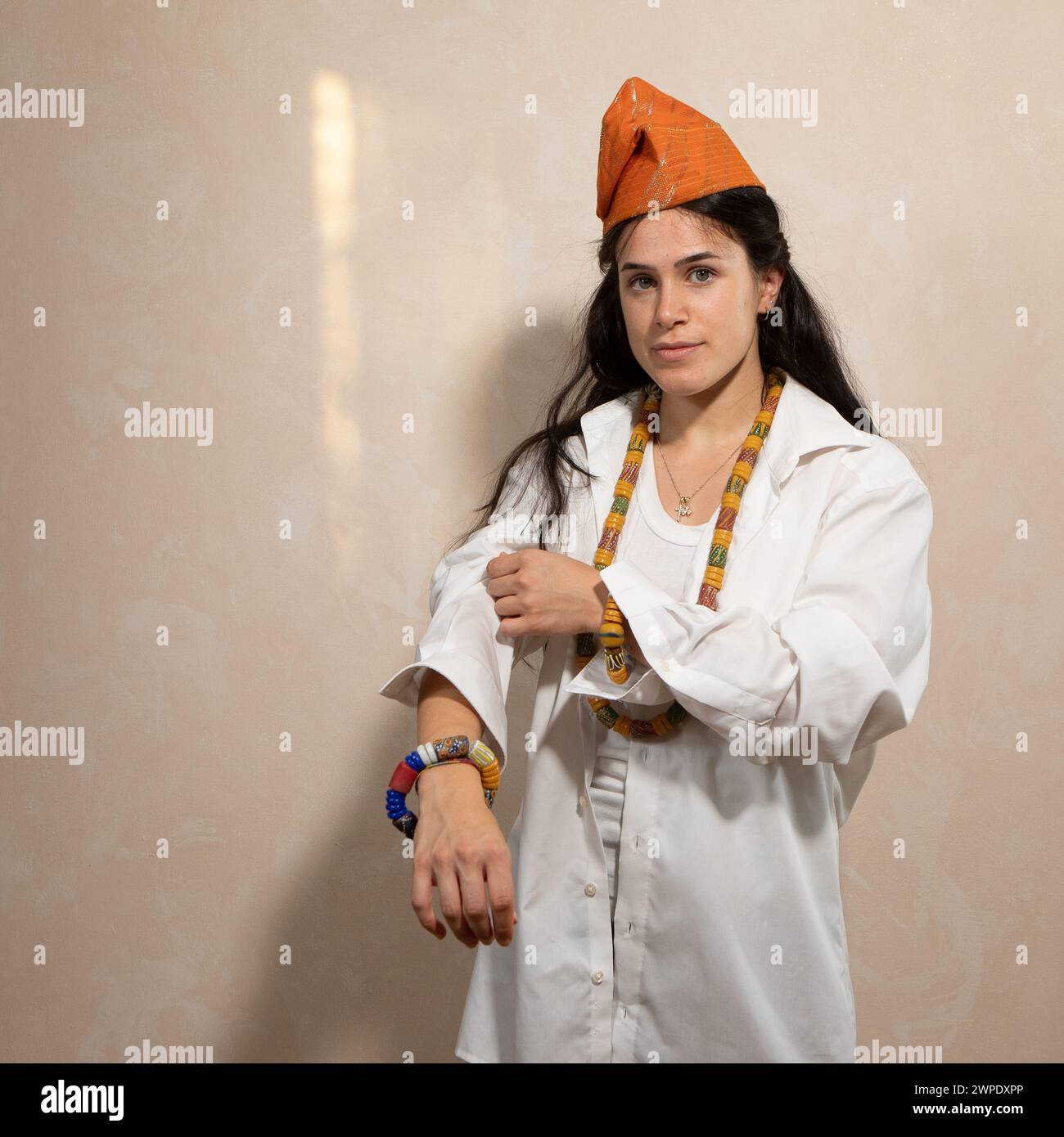 Une jeune femme confiante dans une chemise blanche croustillante et un chapeau africain traditionnel se tient debout, orné de perles colorées, incarnant un mélange de sty moderne Banque D'Images