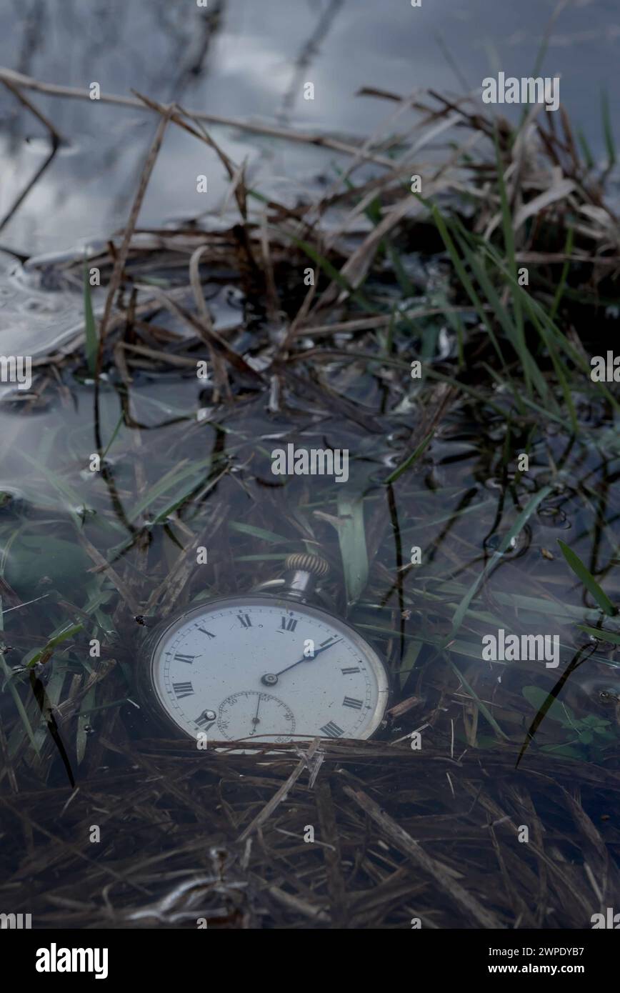 Une vieille montre de poche argentée posée dans une eau peu profonde sur le bord d'un étang, des nuages se reflétant dans l'eau. Banque D'Images