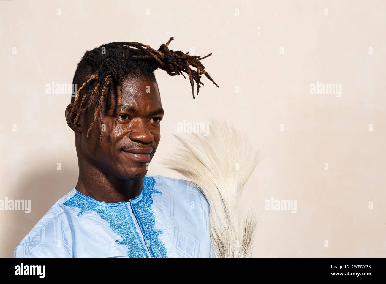 Photo de profil d'un Africain confiant portant une tenue traditionnelle bleu clair avec broderie détaillée, tenant un éventail de crins de cheval. Son unique tordu Banque D'Images