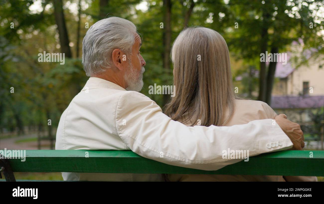 Vue arrière mignon positif des années 70 couple âgé retraité famille heureuse se relaxant en plein air parc naturel de la ville banc. Époux marié homme caucasien femme embrassant Banque D'Images