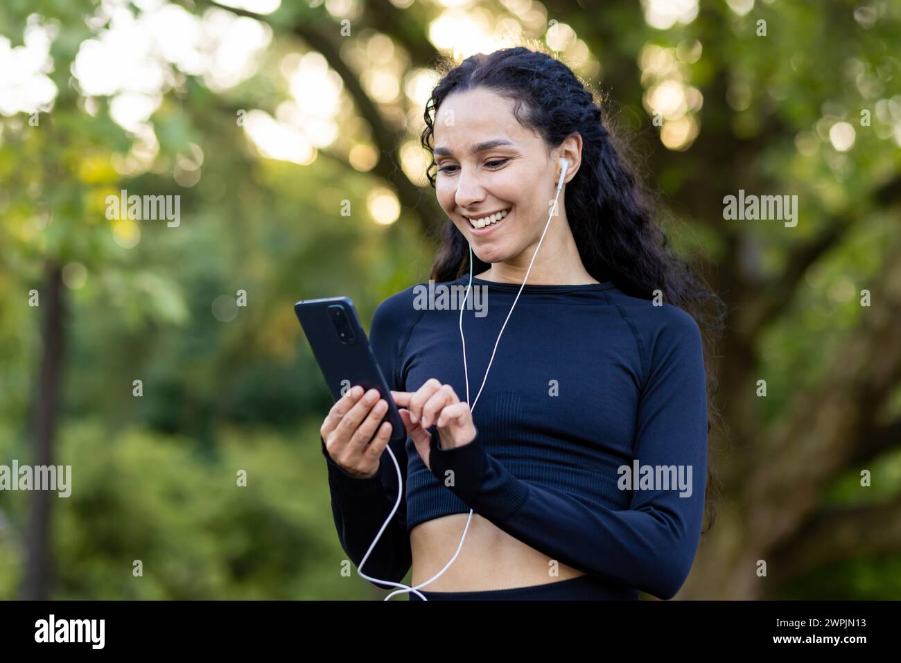 Joyeuse jeune femme active avec des écouteurs souriant tout en tenant un téléphone, profitant d'une pause dans un parc luxuriant. Banque D'Images