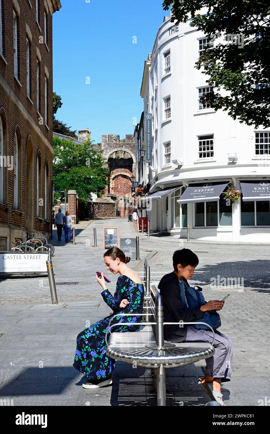 Les gens assis sur des bancs utilisant des téléphones mobiles avec vue le long de Castle Street vers le château de Rougemont (également connu sous le nom de château d'Exeter, Exeter, Devon, Royaume-Uni. Banque D'Images
