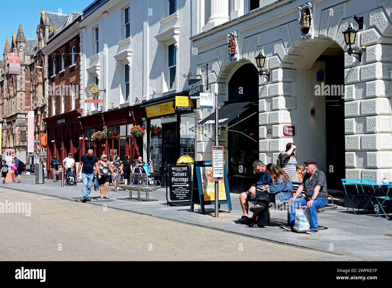 Les gens assis sur un banc près d'un arrêt de bus avec des magasins et des acheteurs le long de Queen Street dans le centre-ville, Exeter, Devon, Royaume-Uni, Europe. Banque D'Images