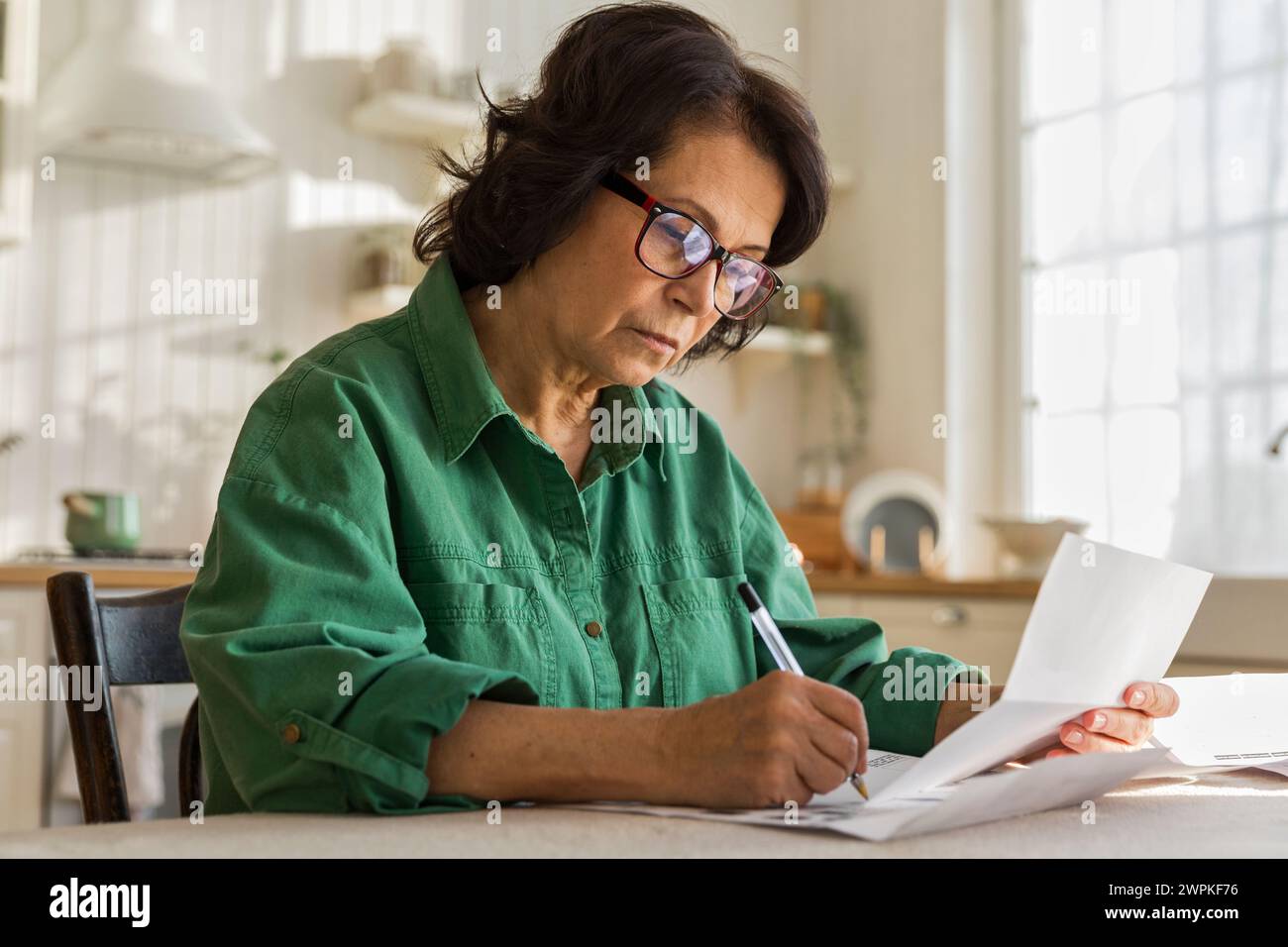 une femme âgée sérieuse travaille avec des papiers et des factures, assise dans la cuisine Banque D'Images