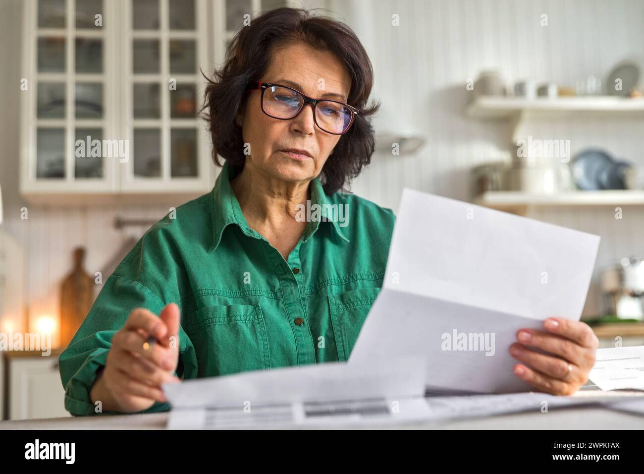 une femme âgée réfléchie lit des papiers et des factures, assise dans la cuisine Banque D'Images