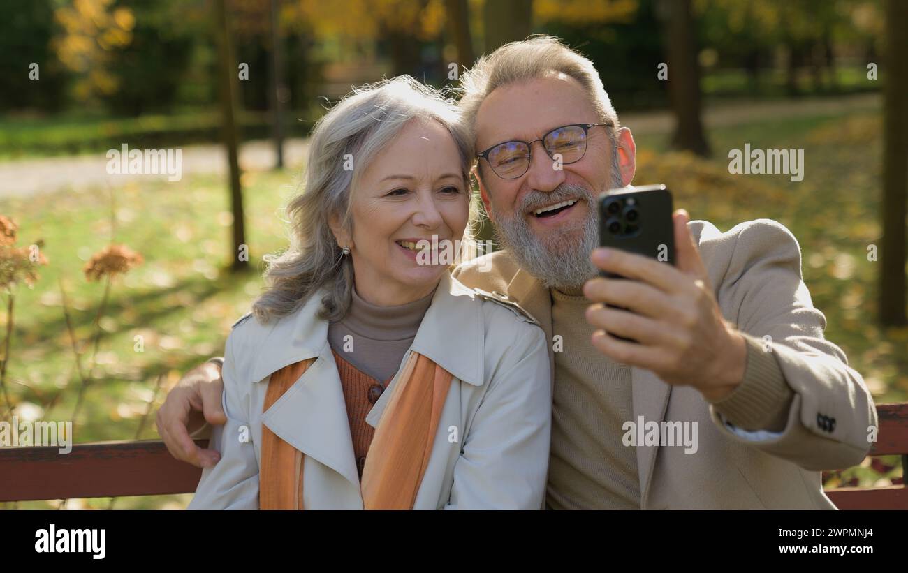 Mari de femme aux cheveux gris joyeux utilisant la communication en ligne de smartphone sur le banc dans la nature du parc d'automne vieux grands-parents couple senior famille mature Banque D'Images