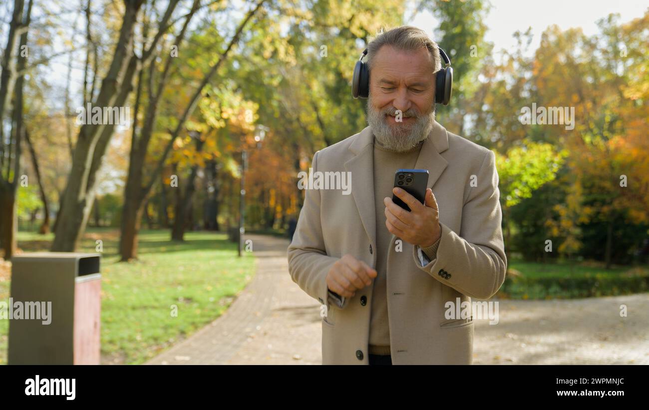 Homme caucasien senior 60s vieux mâle marcher dans la ville de parc d'automne écouter de la musique dans des écouteurs sans fil dansant regarder la vidéo musicale sur téléphone mobile heureux Banque D'Images