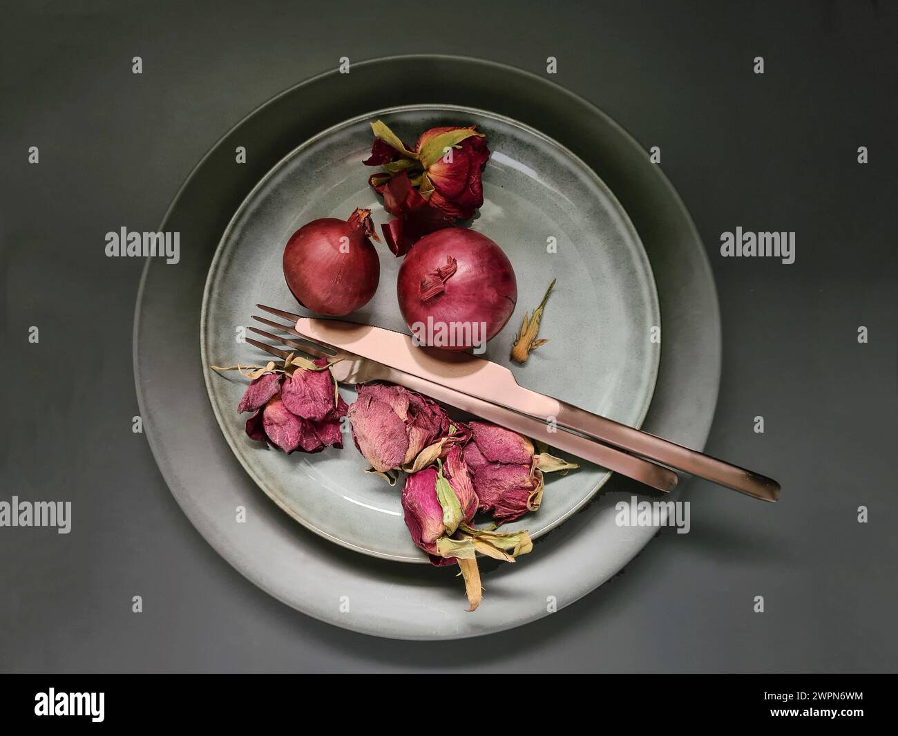 Arrangement de deux oignons rouges entiers avec des pétales de rose séchés à côté de couverts cuivrés sur une assiette en porcelaine comme une nature morte Banque D'Images