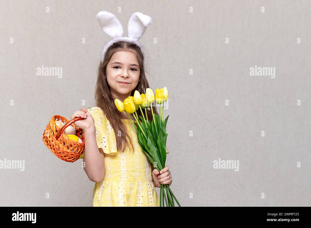 Une fille avec des oreilles de lapin sur la tête avec un panier d'oeufs colorés et des fleurs jaunes dans ses mains. Pâques pour un enfant. Banque D'Images