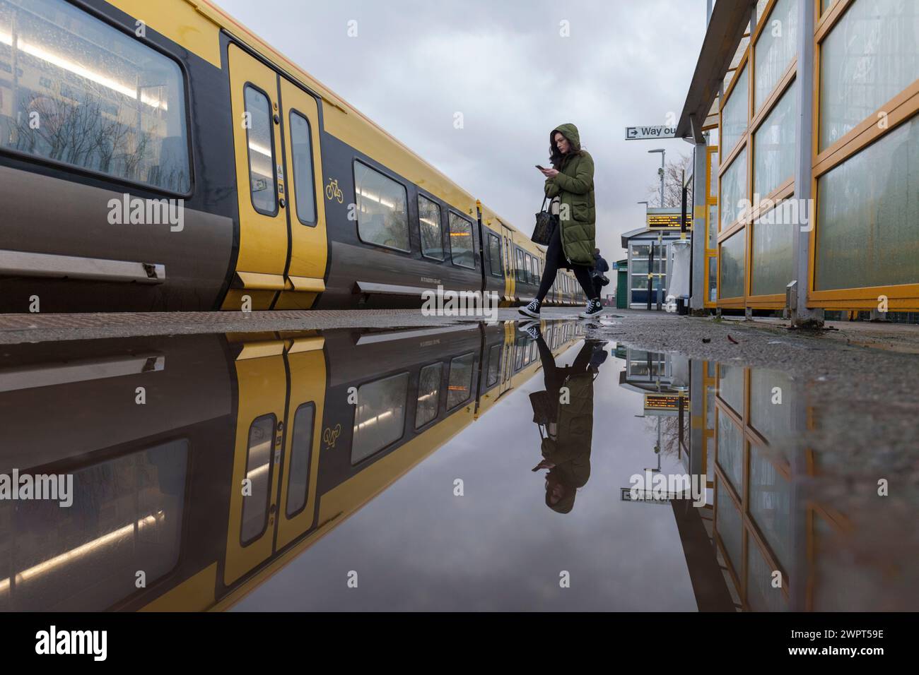 Passager embarquant dans un train Merseyrail Stadler classe 777 à la gare de Bebington reflété dans une flaque sur le quai de la gare Banque D'Images
