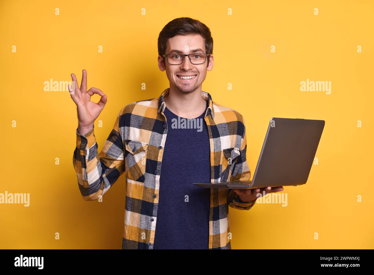 Portrait de l'homme cool intelligent et confiant positif dans des lunettes tenant un ordinateur portable, faire un geste de main correct isolé sur fond jaune, trouver de nouveaux clients sur les médias sociaux, l'apprentissage en ligne, la technologie Banque D'Images