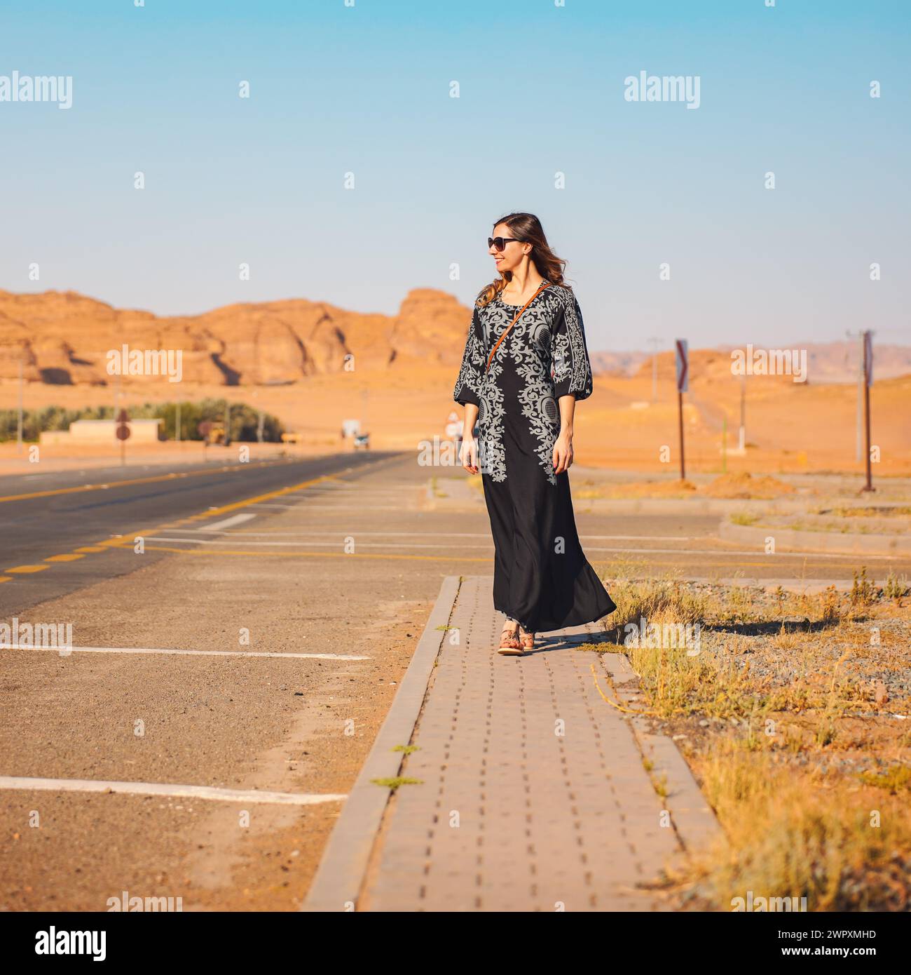 Jeune femme en longue robe noire marchant sur le trottoir près de la route - paysage rocheux désert, typique pour Alula, en arrière-plan Banque D'Images