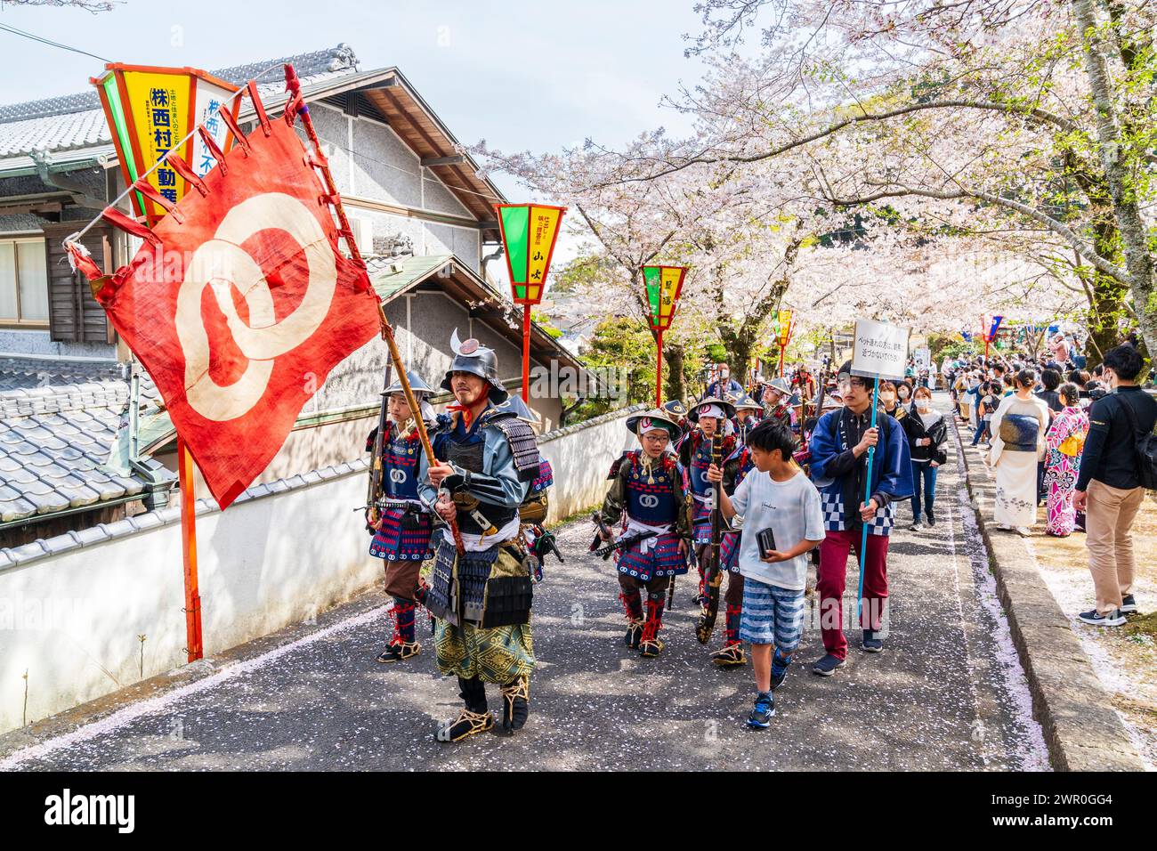 Défilent des samouraïs Tatsuno sous des cerisiers en fleurs avec une bannière portant des samouraïs menant un groupe de garçons habillés en Teppou ashigaru. Les gens regardent. Banque D'Images