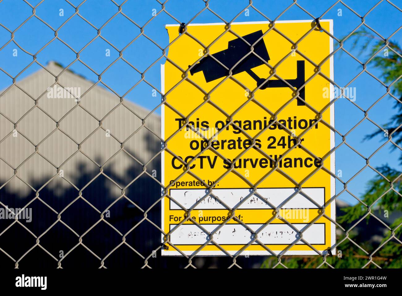 Gros plan d'un panneau jaune attaché à une clôture à maillons de chaîne avertissant que la vidéosurveillance fonctionne 24 heures sur 24 dans les locaux d'une entreprise industrielle. Banque D'Images