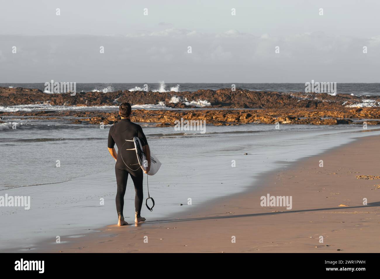 Surfeur en combinaison debout seul sur le sable regardant la mer avec sa planche de surf sous le bras Banque D'Images
