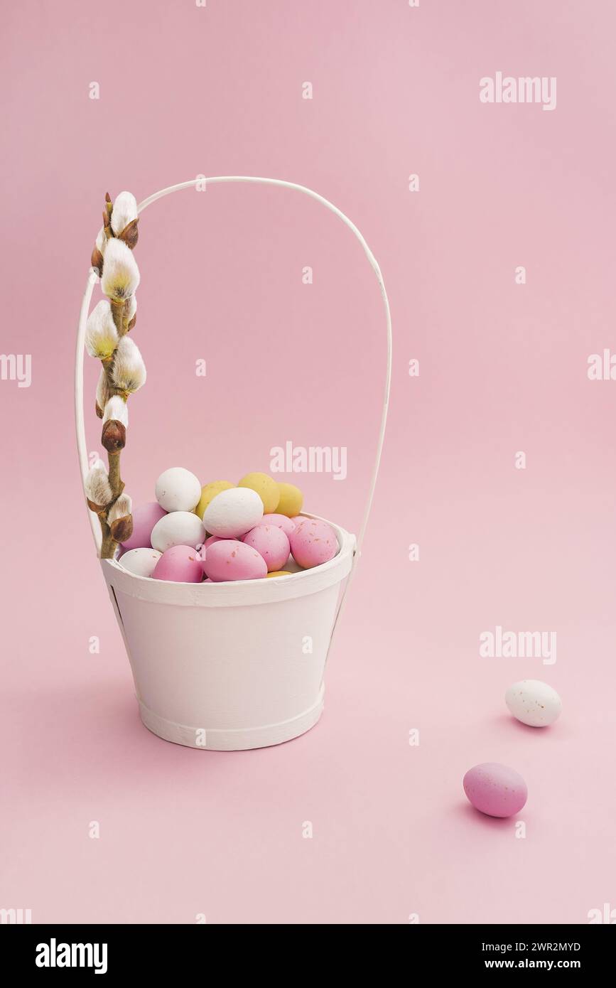 Panier de Pâques blanc avec œufs multicolores en chocolat et branche de saule sur fond rose pastel. Composition de Pâques. Carte avec espace de copie pour le texte Banque D'Images