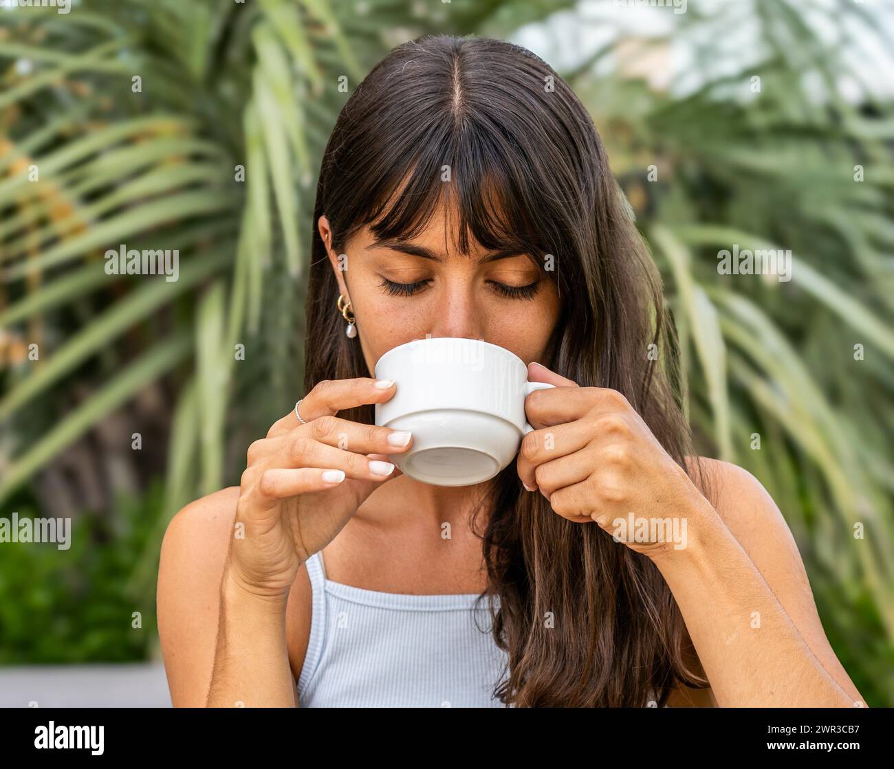 Une femme boit du café dans une tasse blanche les yeux fermés. Elle porte un débardeur et a ses cheveux tirés en arrière Banque D'Images
