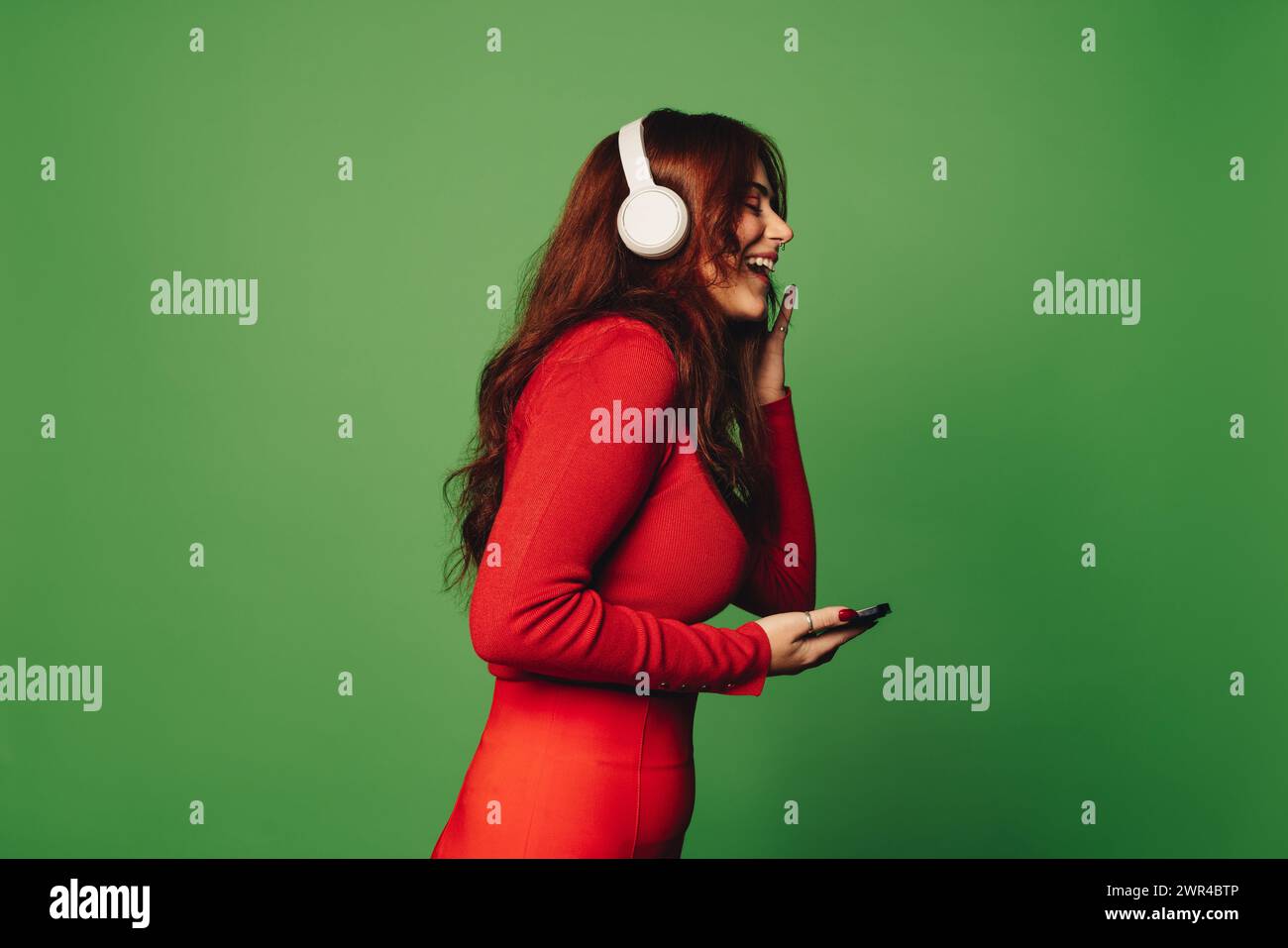 Jeune femme dans des vêtements décontractés se tient sur un fond vert, souriant et tenant un smartphone avec des écouteurs. Heureuse jeune femme appréciant le Banque D'Images