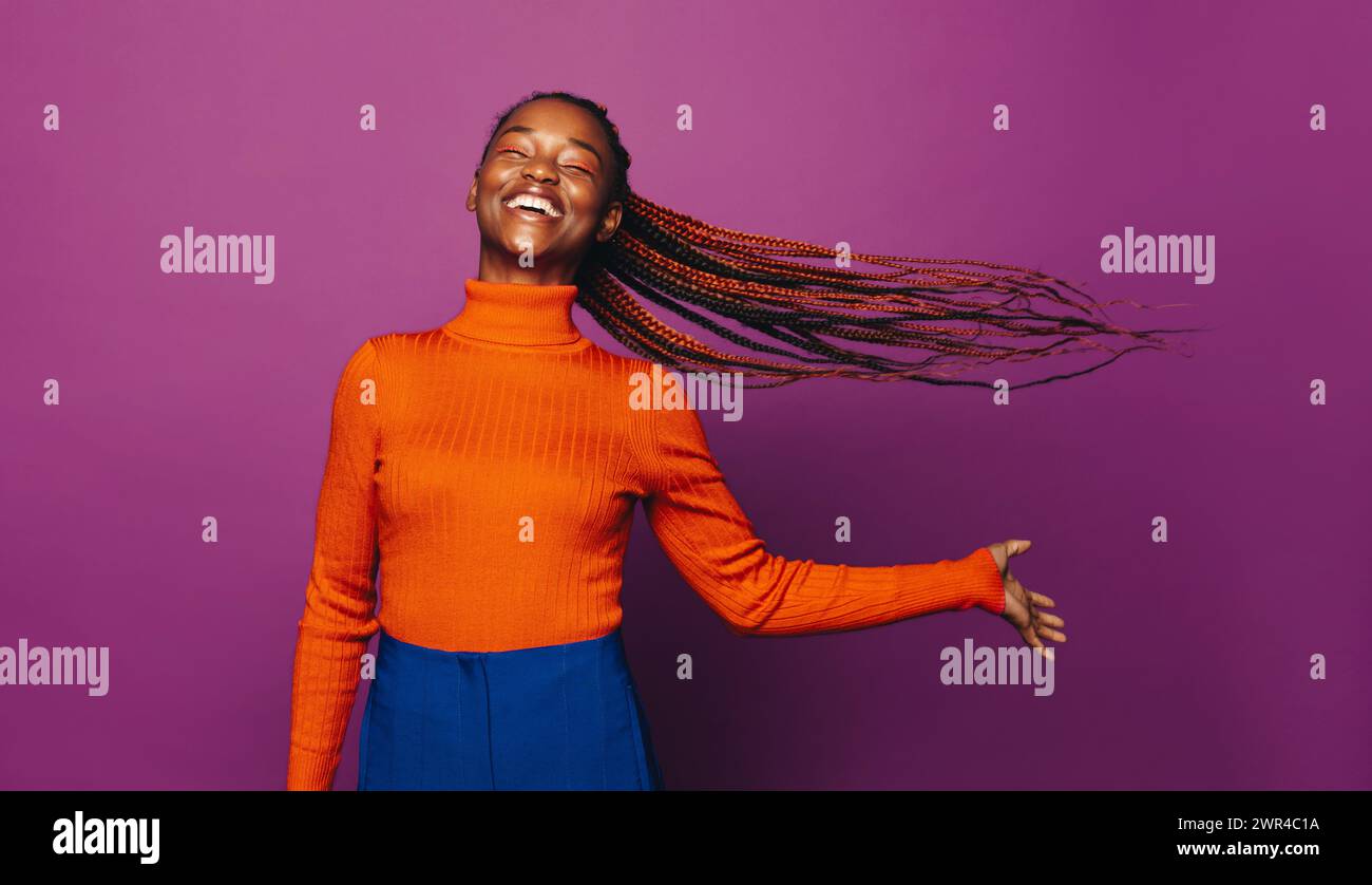 Femme africaine dans la vingtaine avec des tresses bicolores élégantes se tient dans un studio, souriante. Elle porte des vêtements décontractés, un Jean et un col polo, célébrant la vie wi Banque D'Images