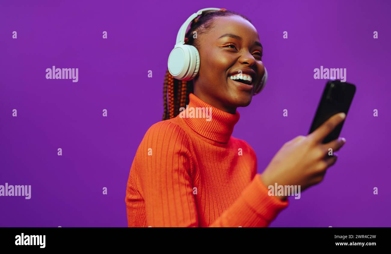 Femme porte des vêtements décontractés et un col polo, stylisé avec des tresses bicolores et un maquillage eyeliner. Elle tient un smartphone avec des écouteurs violets, navigue Banque D'Images