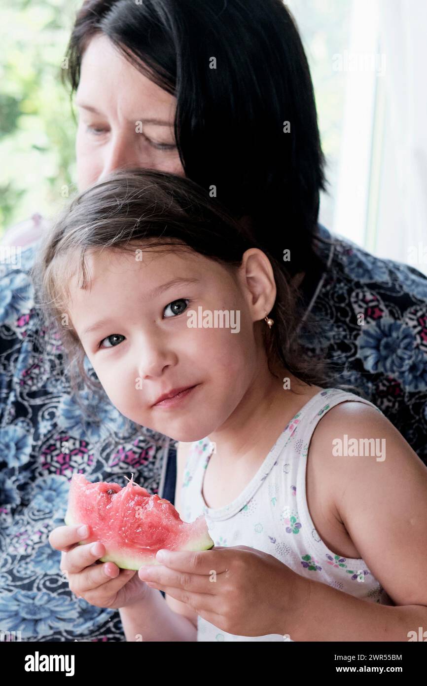 Mignonne petite fille assise près de maman mangeant un morceau de pastèque et regardant la caméra. Banque D'Images