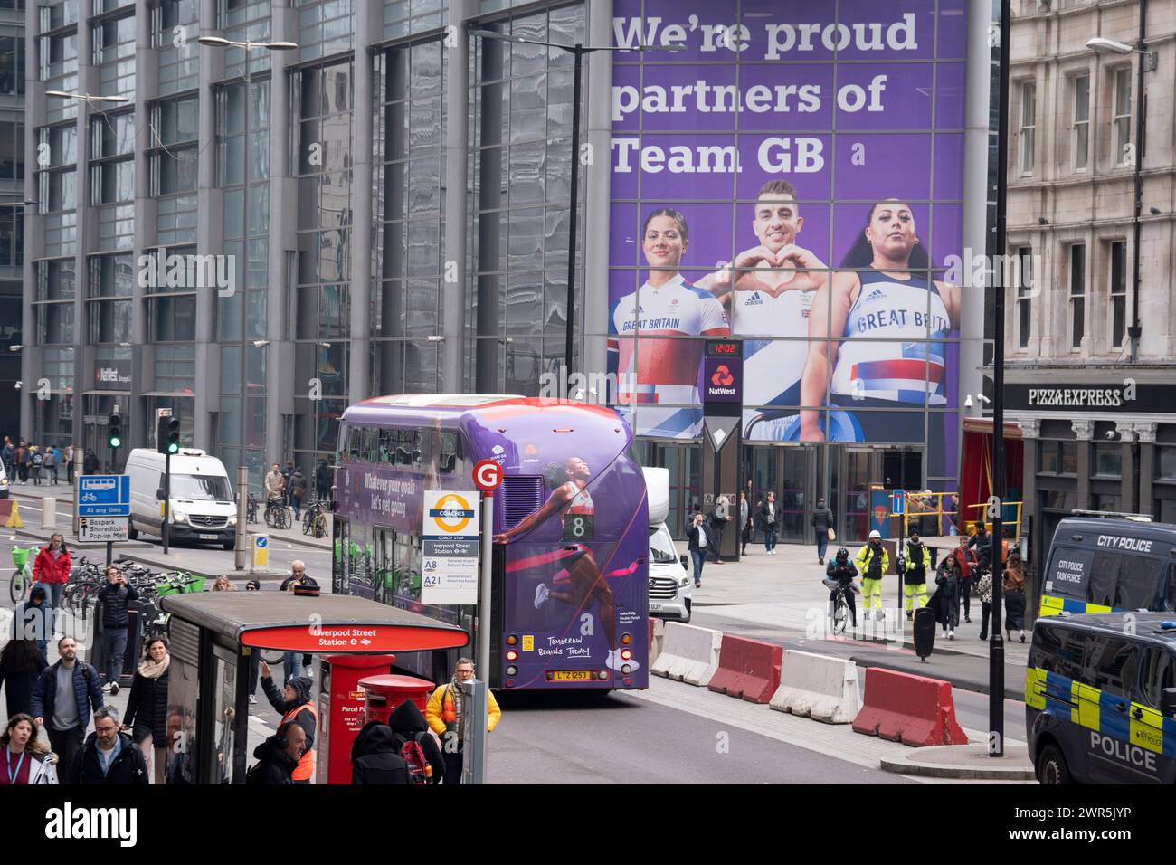 Quelques mois avant les Jeux olympiques de Paris en 2024, un bus londonien portant une image de la sprinteuse Asha Philip passe devant une énorme image d'autres athlètes de l'équipe GB, la coureuse BMX Beth Shriever, le gymnaste Max Whitlock et l'haltérophile Emily Campbell à l'entrée de Natwest Bank sur Bishopsgate dans la ville de Londres, le quartier financier de la capitale, le 7 mars 2024, à Londres, Angleterre. NatWest et Coutts Bank sont actuellement les partenaires officiels de Team GB. Banque D'Images