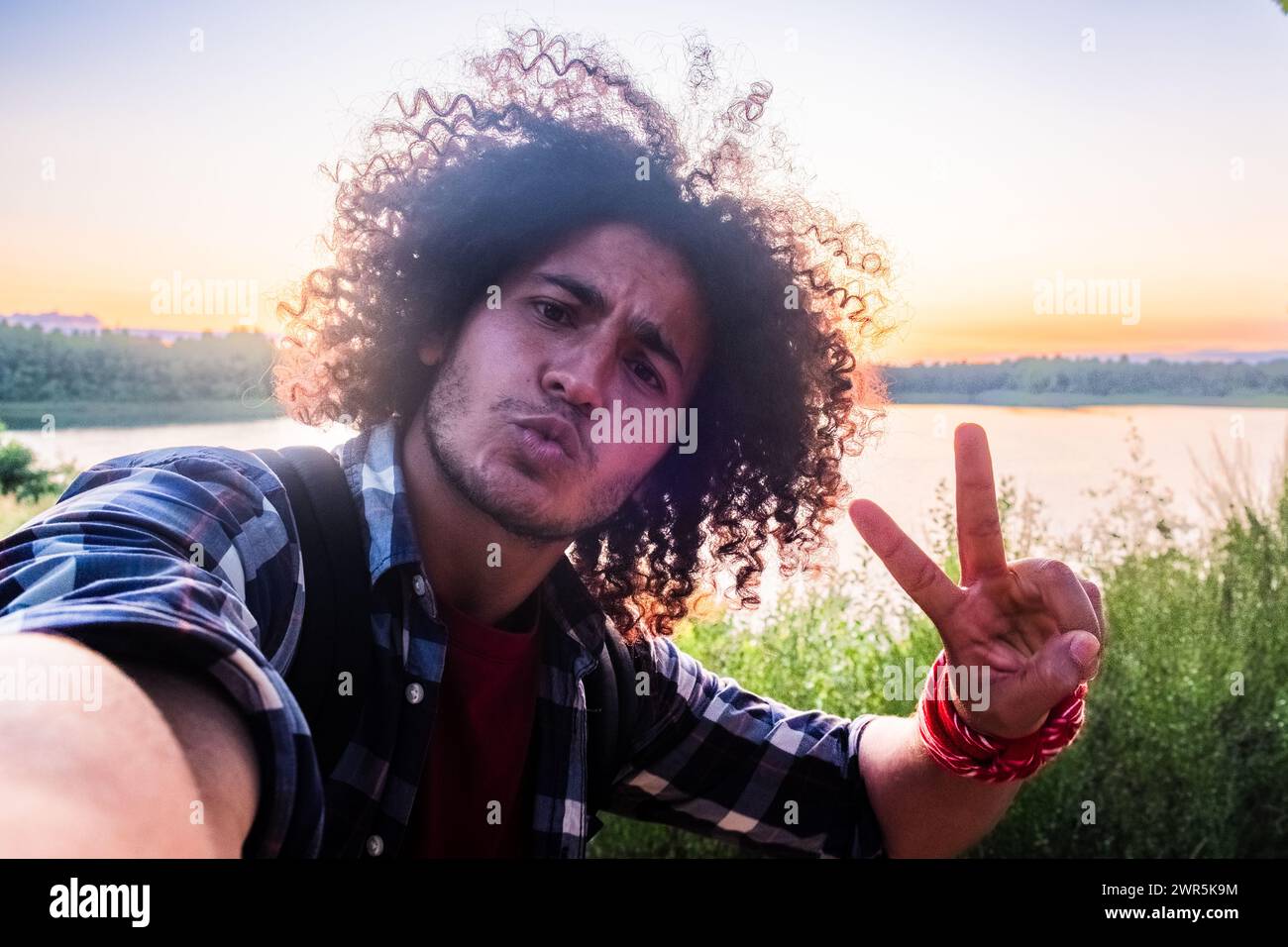 Jeune adulte, cheveux bouclés, donnant signe de paix, à l'extérieur, coucher de soleil en toile de fond, expression franche. Homme aux cheveux bouclés faisant signe de paix pendant le Selfie au coucher du soleil. Photo de haute qualité Banque D'Images