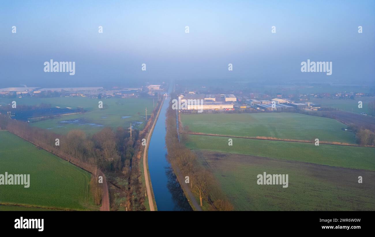 Une scène matinale brumeuse se déroule, révélant la juxtaposition des champs agricoles et du développement industriel par le haut. Vue surélevée du canal brumeux divisant les terres agricoles rurales et la zone industrielle. Photo de haute qualité Banque D'Images