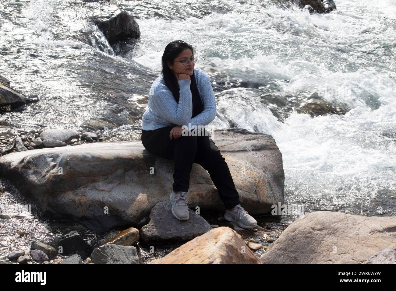 Femme souriante est assise tranquillement sur un rocher surdimensionné, la main sur le menton, près des eaux jaillissantes du fleuve Gange à Rishikesh Banque D'Images