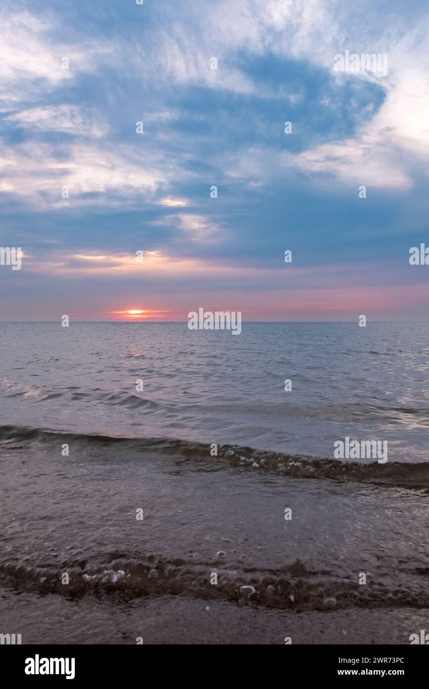 Cette photographie capture la majesté tranquille d’un coucher de soleil sur la mer, avec le soleil plongeant sous l’horizon, sa lumière diffusée par un voile de nuages. Le ciel est une toile de bleus doux et de roses subtils, reflétant le salut final du soleil à la journée. La mer, douce et tamisée, chevauche le rivage rocheux, contribuant à l'atmosphère sereine. Les nuages sont artistiquement striés à travers le ciel, ajoutant de la profondeur et de la texture à l'affichage céleste. Cette scène évoque un sentiment de calme et d’introspection alors que le jour passe à la nuit. Le délice du crépuscule : coucher de soleil tranquille sur la mer douce. Photo de haute qualité Banque D'Images