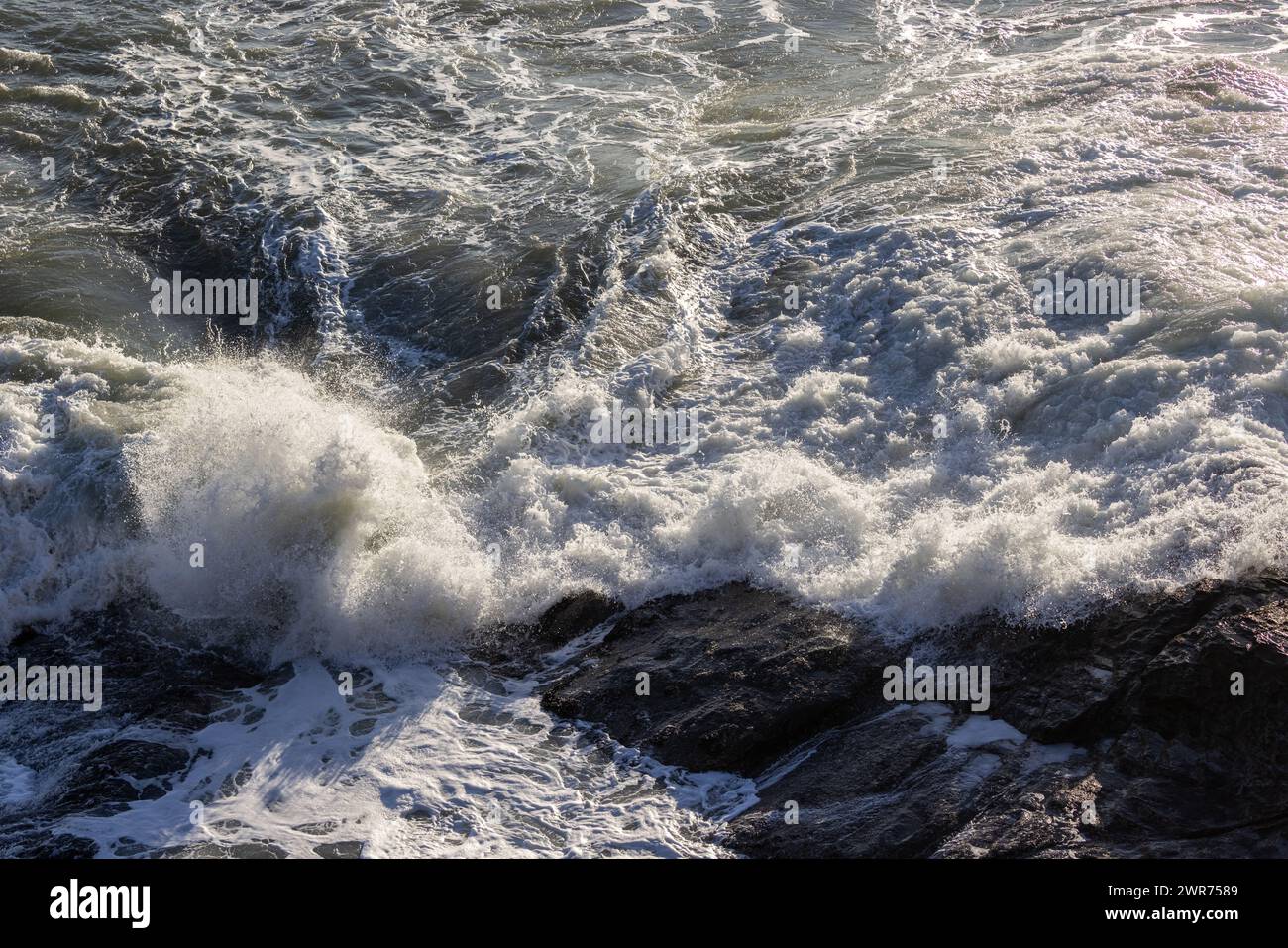 Une grosse vague s'écrasant contre un rivage rocheux. L'eau est blanche et mousseuse, et les rochers sont dispersés dans toute la scène. La scène est puissante et le dr. Banque D'Images