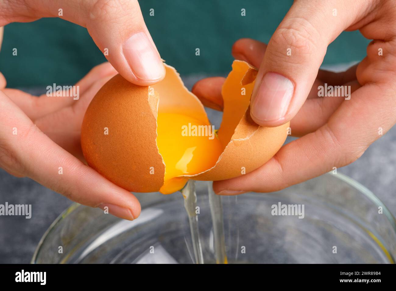 Les mains de femme au foyer craquant l'œuf frais, le jaune et le blanc tombant dans un bol. La main d'une femme cassant un oeuf avec un couteau dans un bol en verre transparent dans le k gris Banque D'Images
