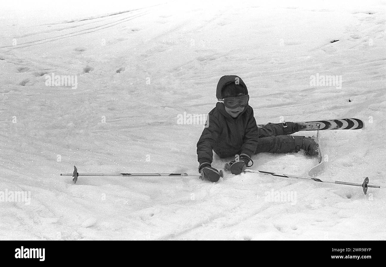années 1970, historique, un jeune garçon, en kit de ski et googles, sur le sol tenant ses skis étant tombé dans ses tentatives d'apprendre à skier. Banque D'Images