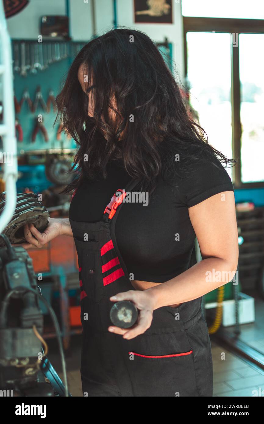 Jeune femme brune hispanique habile confiante aux cheveux longs dans le tablier d'un mécanicien examine une pièce de voiture dans un atelier, un panneau d'outils complet dans Banque D'Images