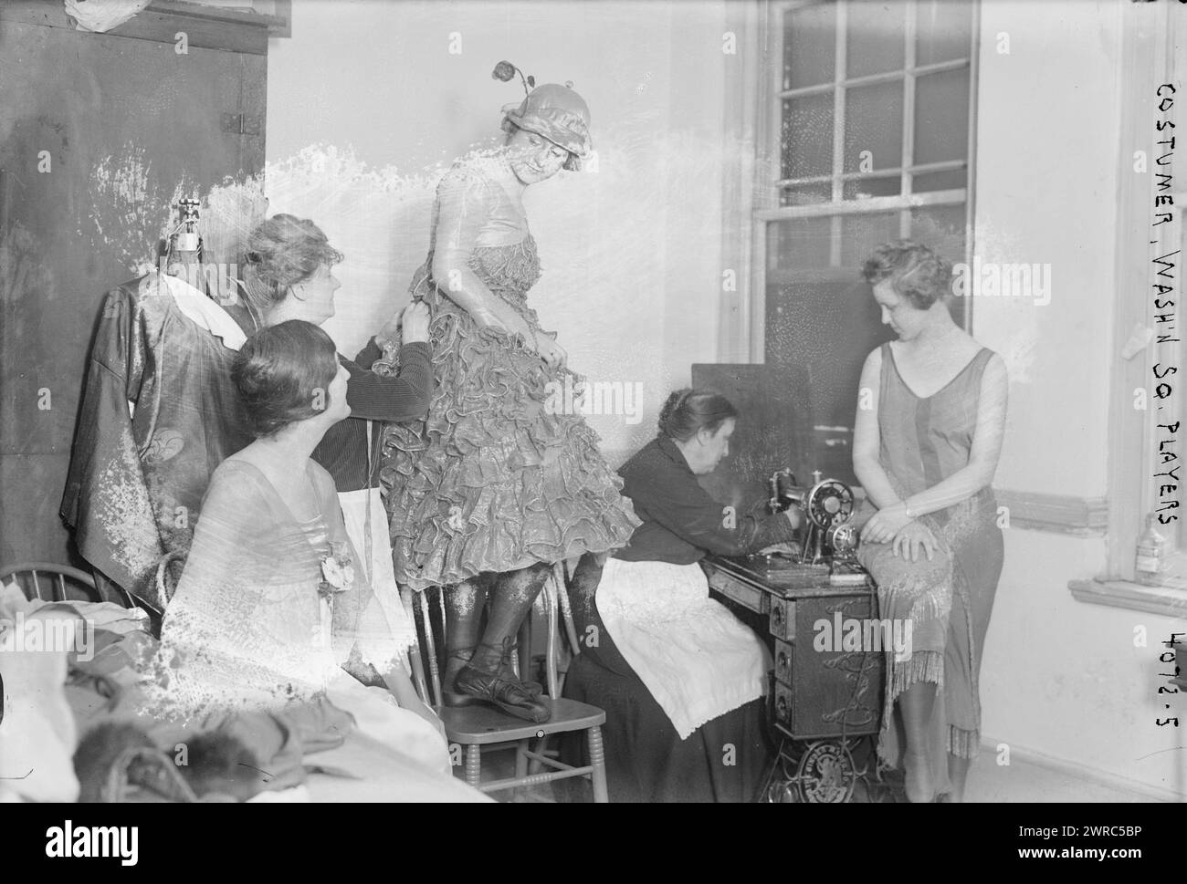 Costumer, Wash'n Sq. C'est-à-dire les joueurs de Washington Square, la photographie montre le costumier à une machine à coudre, préparant des costumes conçus par George Wolfe Plank (1883-1965) pour la première production des joueurs de Washington Square de la saison 1916, 'A Merry Death' de Nicholas Evreinov. Florence Enright habillée en Columbine est debout sur une chaise derrière le costumer., 1916, négatifs en verre, 1 négatif : verre Banque D'Images