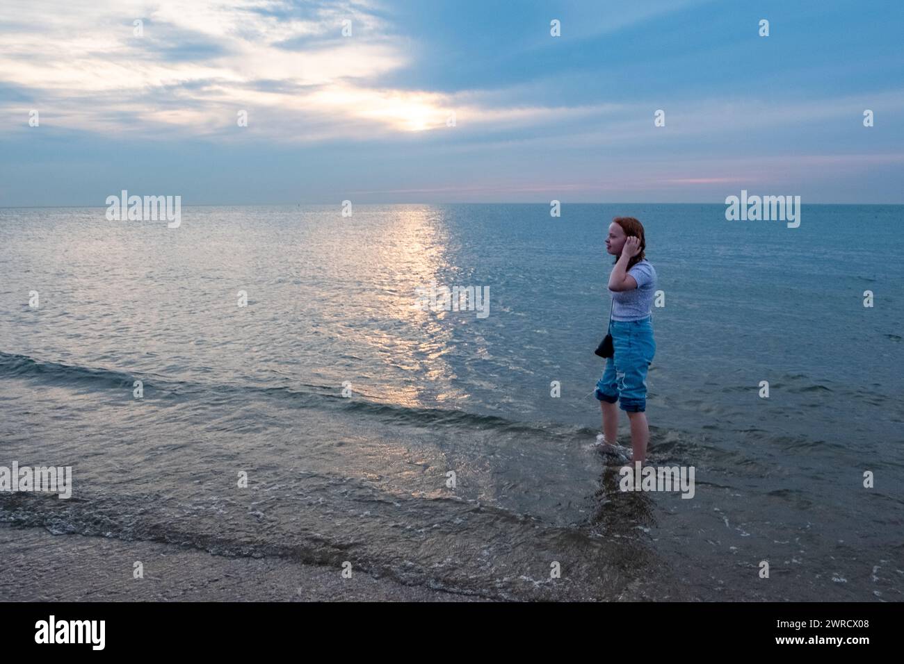 Cette image représente une jeune personne debout dans les eaux peu profondes de la mer, avec ses mains doucement placées sur ses oreilles, comme pour mieux entendre le murmure des vagues. Ils regardent vers le coucher du soleil, délicatement voilé par les nuages, projetant un doux reflet sur la surface de l'eau. Le Jean décontracté et retroussé et le t-shirt ajusté suggèrent une fin de journée décontractée, mais introspective. Le calme de la mer et le ciel pastel contribuent au sentiment général de paix et de solitude. Jeune individu embrassant Sea Air au coucher du soleil. Photo de haute qualité Banque D'Images