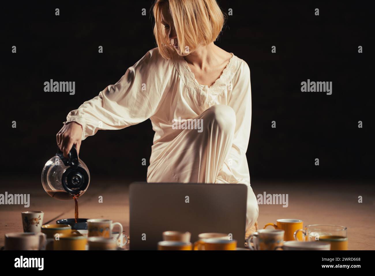 Recyclage, travail acharné de freelance, concept d'échéance. Une femme dans une chemise blanche verse du café dans de nombreuses tasses devant un ordinateur portable. Une pièce sombre. Banque D'Images