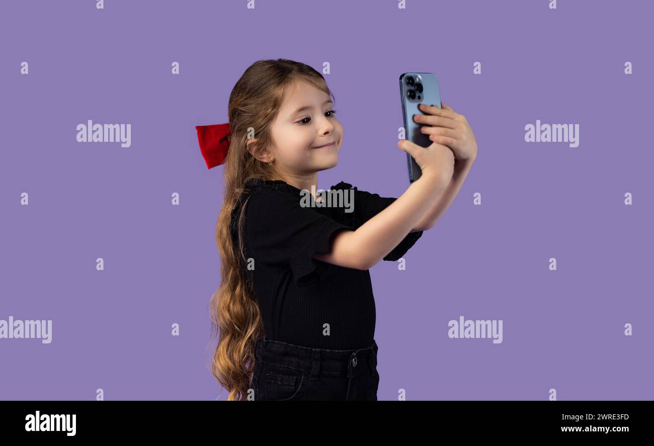 Enfants et technologie. La jeune influenceuse joyeuse à la mode enregistre un vlog vidéo et prend un selfie sur fond violet. Drôle de fille preteen mignonne Banque D'Images