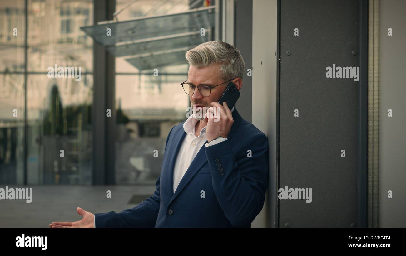 Homme inquiet caucasien en colère des années 50 argumente parler smartphone fou dans la ville penchant mur de l'immeuble de bureaux de la société stressé vieil homme d'affaires répondre à l'appel Banque D'Images