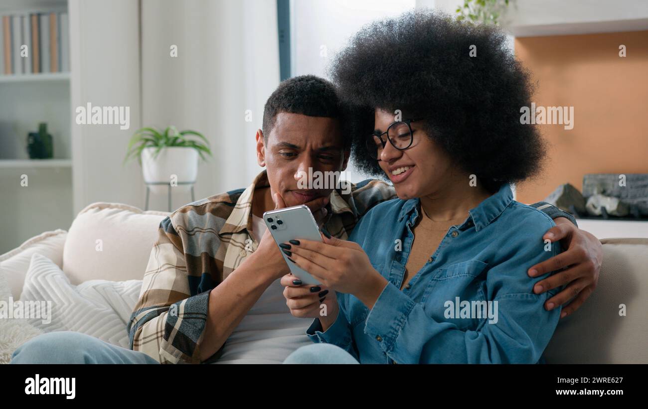 Afro-américain époux couple homme femme fille Guy sur canapé canapé à la maison parler ensemble discuter des achats de famille en ligne avec le téléphone mobile choisir Banque D'Images