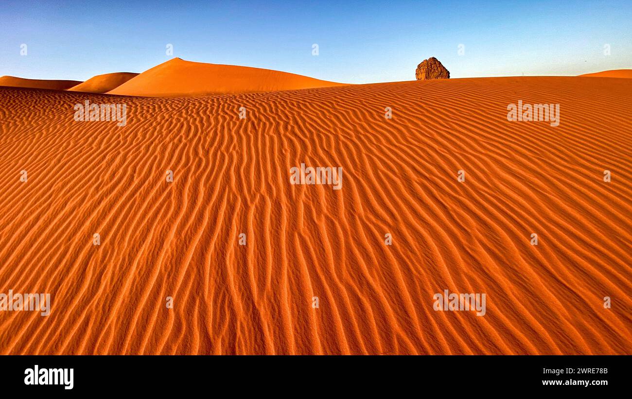 Paysage du Tadrart Rouge dans le désert du Sahara, Algérie. Un rocher émerge des dunes. Le vent façonna les ondulations dans le sable Banque D'Images