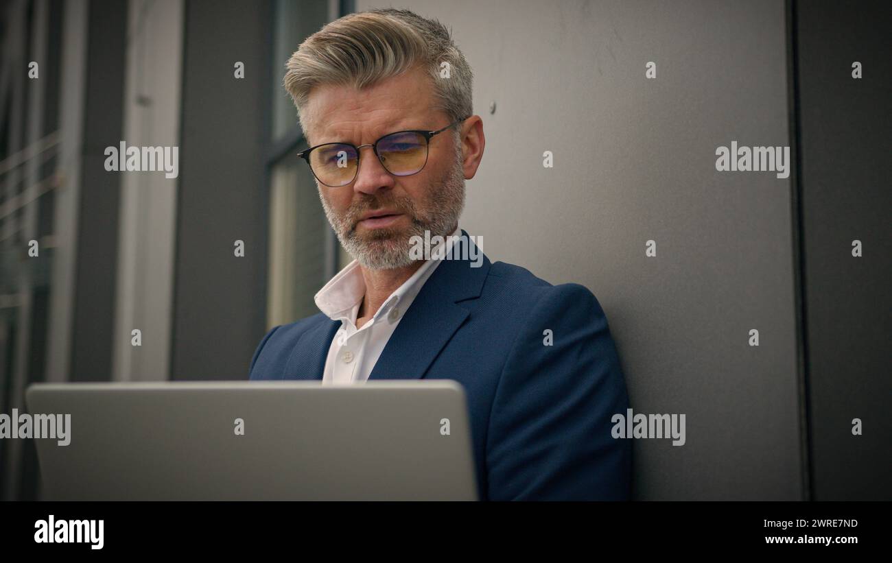 Caucasien âgé homme senior gestionnaire d'affaires mature occupé homme d'affaires d'employeur d'âge moyen penchant bâtiment de bureau de mur tapant ordinateur de travail portable Banque D'Images