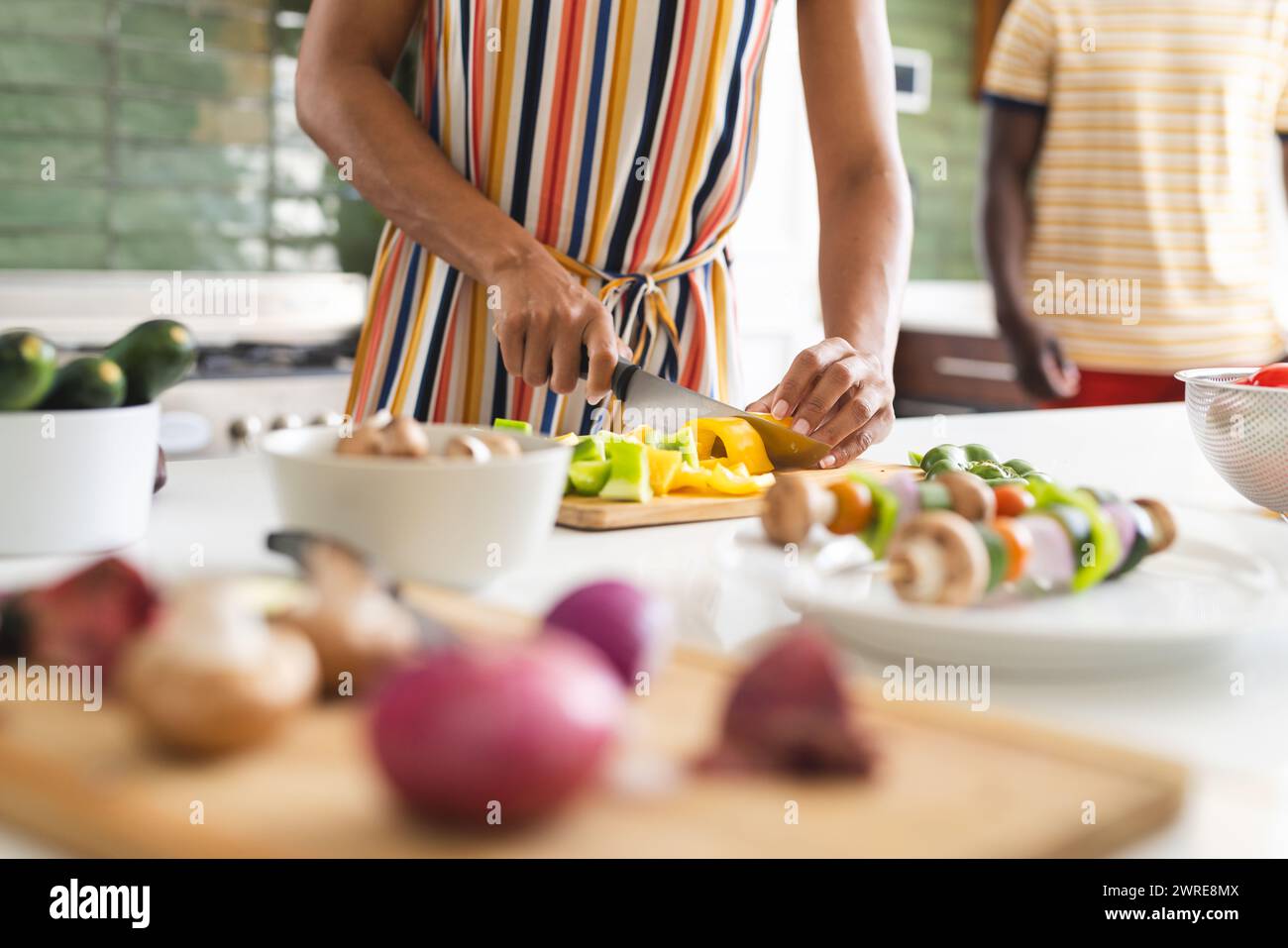 Une femme biraciale dans une robe rayée coupe des légumes dans une cuisine Banque D'Images